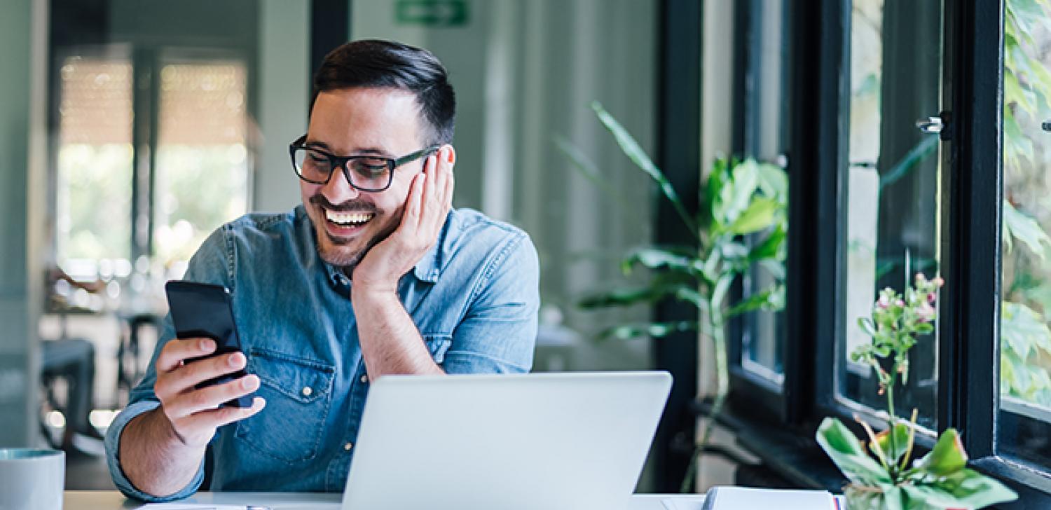 Small Business owners looks at mobile phone smiling while sitting in front of a computer at a desk.