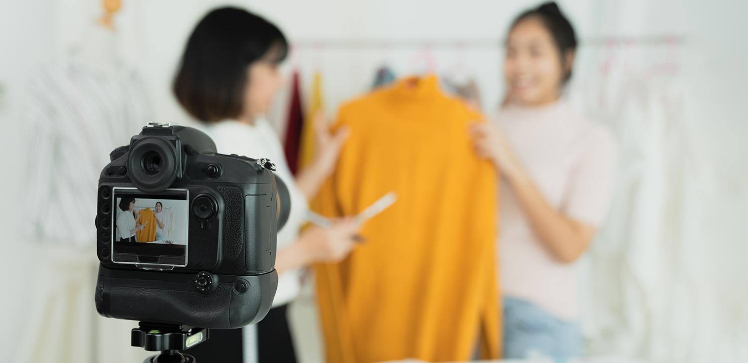 Two women hold up a sweater in front of a camera.